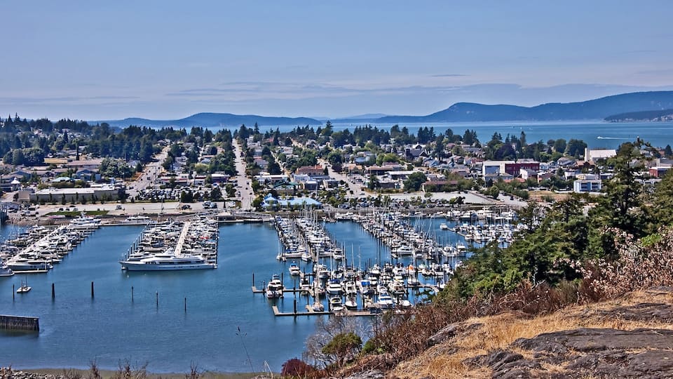A marina with numerous boats docked, surrounded by a coastal town and hills in the background.