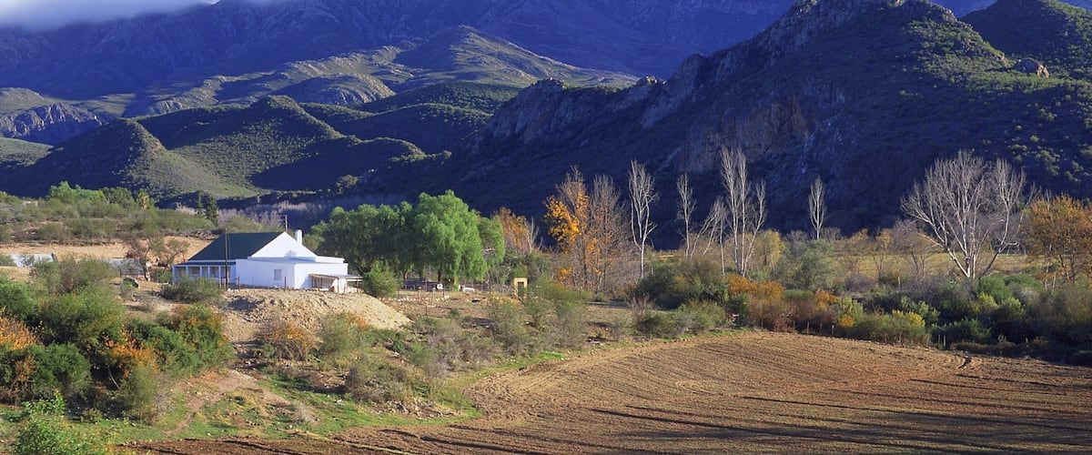 A rural landscape with a white house, rolling hills, and a mountain range in the background.