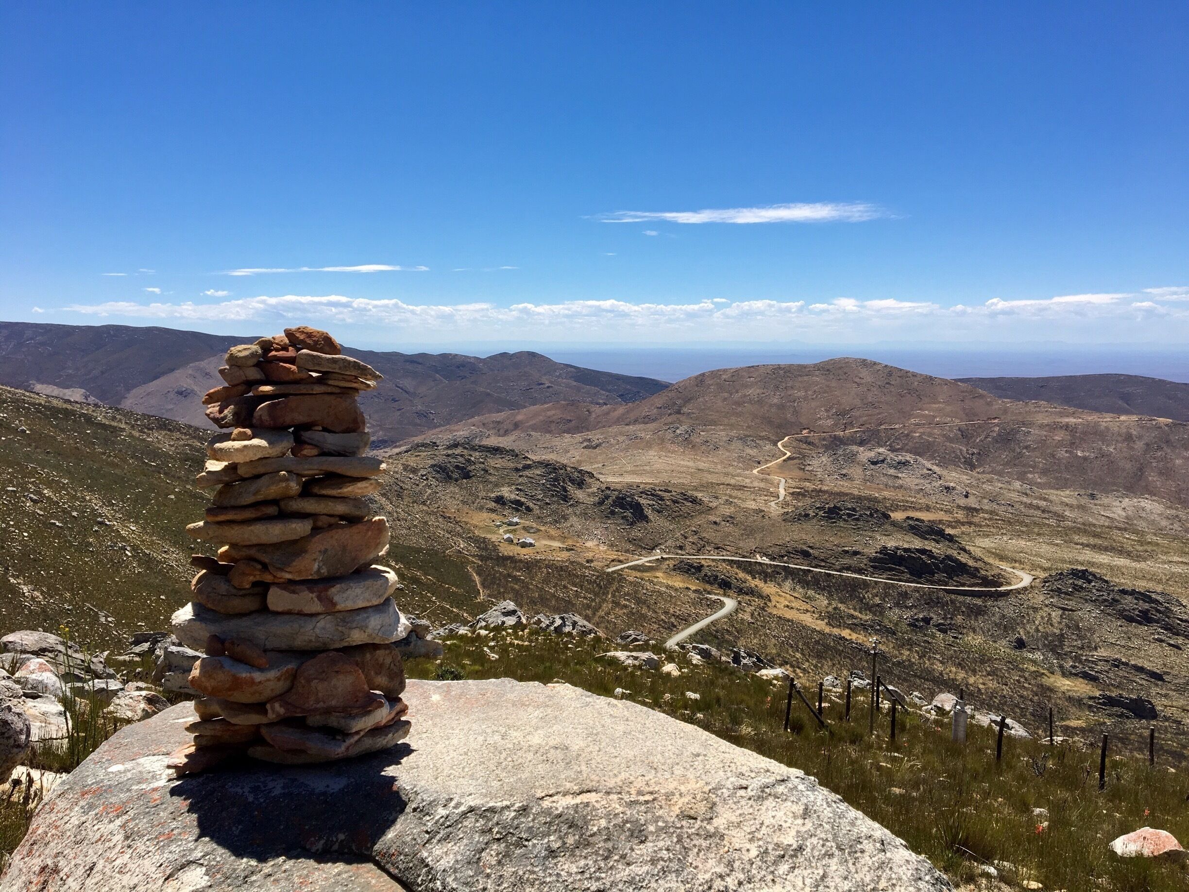 A stone cairn on a mountain ridge overlooking a winding road and distant hills.