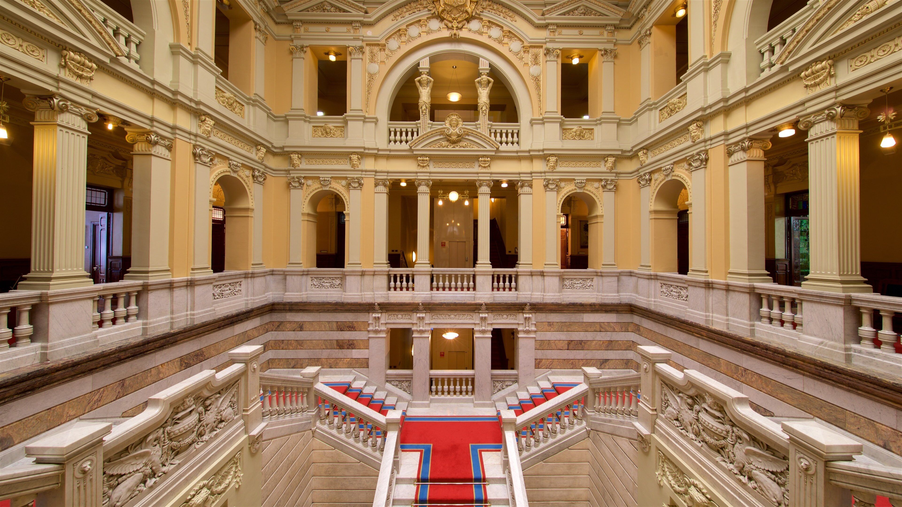 A grand, ornate building with multiple levels, large columns, and a central staircase with red carpeting.