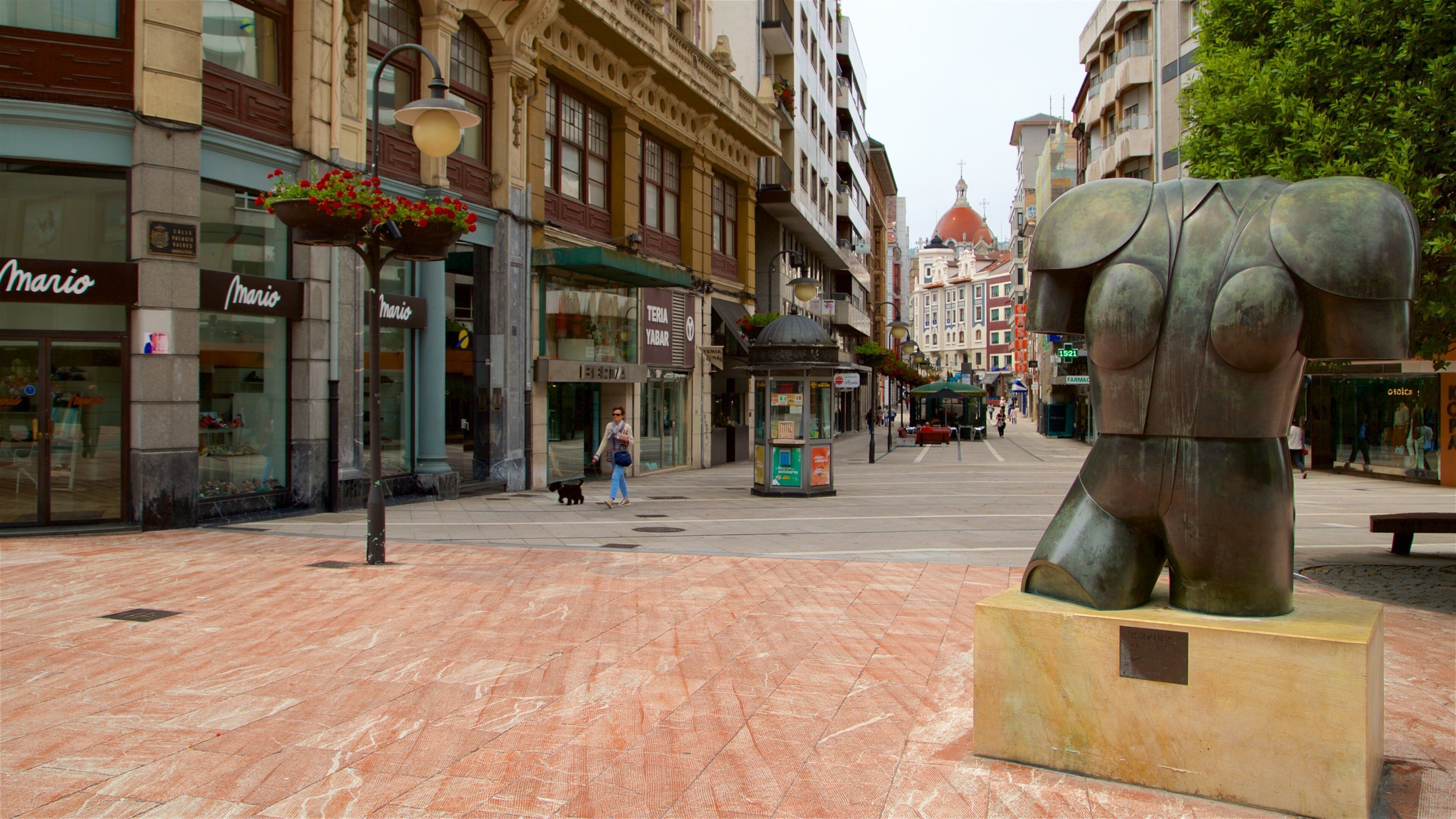 Una plaza con una gran escultura de bronce que representa el torso de una mujer, tiendas y una fuente adornada con flores rojas.