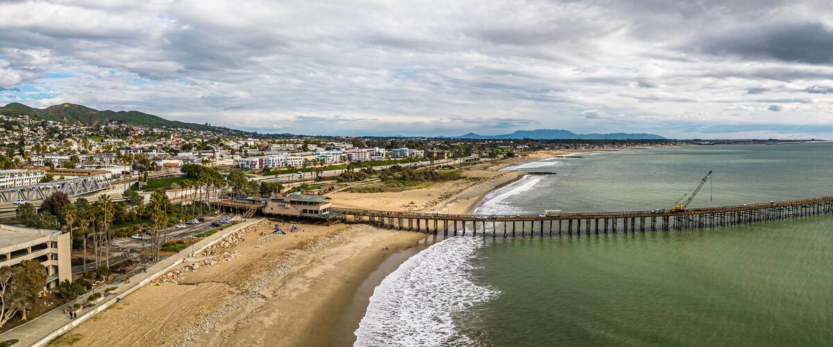 A coastal city with a long pier extending into the ocean, a sandy beach, and a mountainous backdrop.