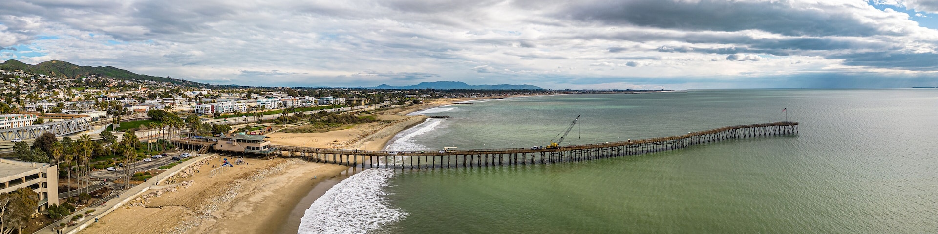 A coastal city with a long pier extending into the ocean, a sandy beach, and a mountainous backdrop.