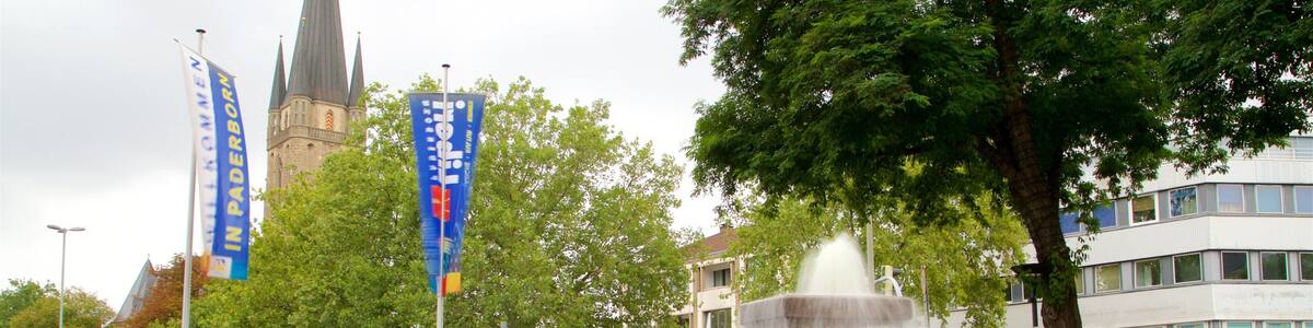 A fountain with multiple tiers, surrounded by a paved area with benches and flower beds. In the background, there are flags, a church tower, and a building with signage.