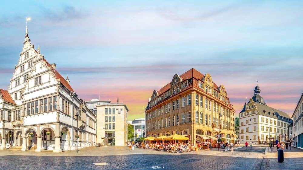 Ein historischer Platz mit einem markanten, weißen Gebäude mit spitzer Dachform, einem großen, gelben Gebäude mit Kuppel und einer gepflasterten Straße.