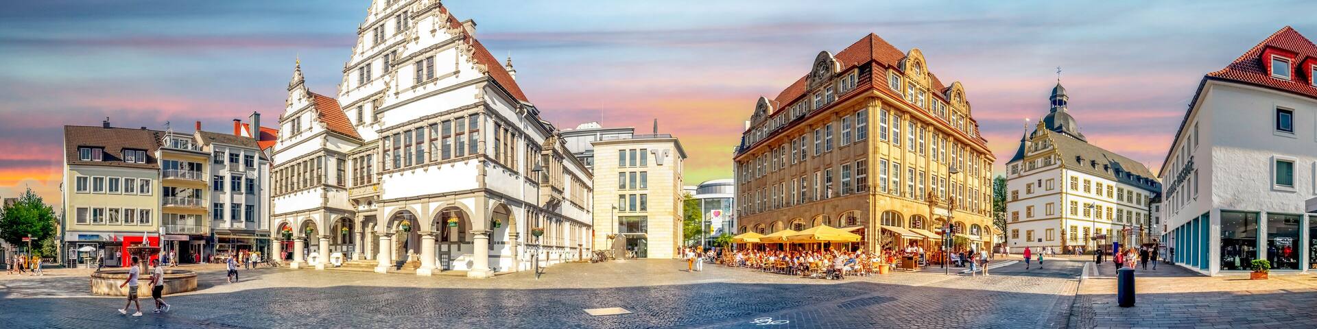 A historic square with a prominent white building with a pointed roof, a large yellow building with a dome, and a cobblestone street.