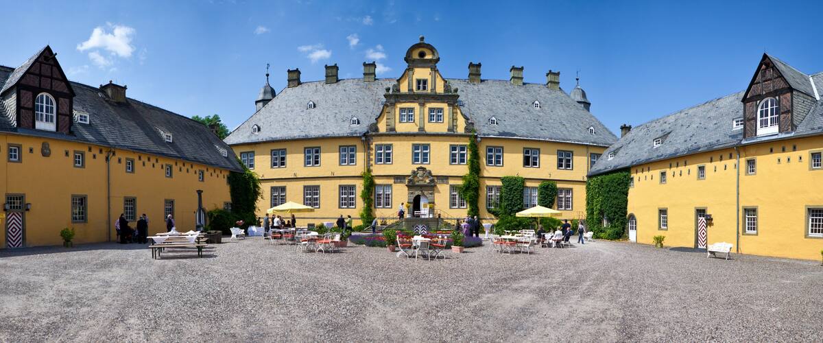 A historic yellow building with a central entrance, surrounded by a gravel courtyard with outdoor seating and people.