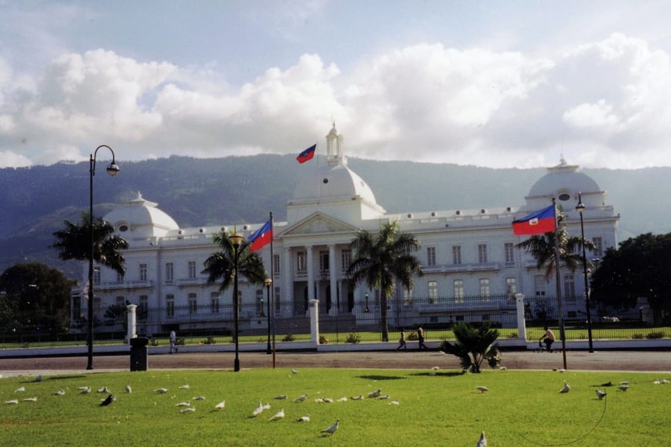 A large white building with domed roofs, surrounded by a well-maintained lawn and palm trees. Flags are flying atop the building.