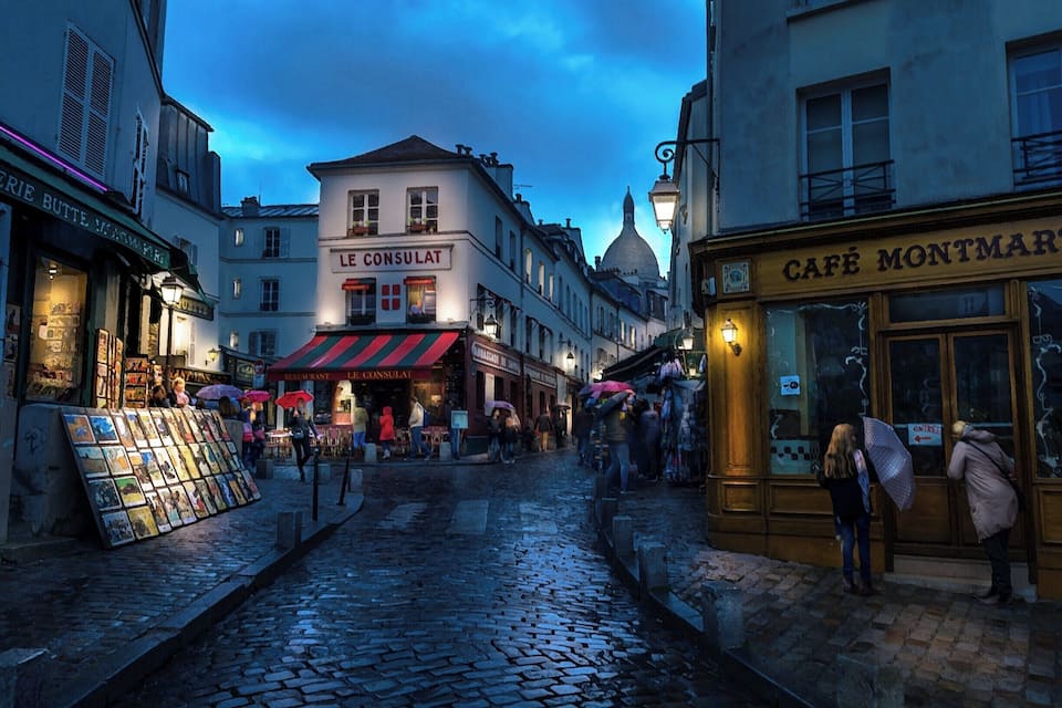 Eine Kopfsteinpflasterstraße mit Geschäften und Cafés, darunter 'Le Consulat' und 'Café Montmartre'.