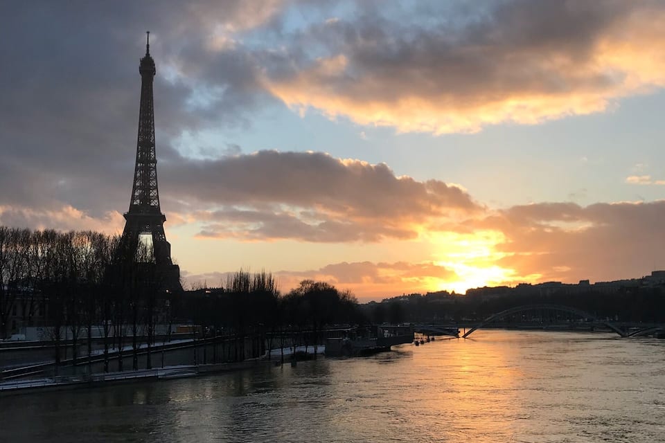 The Eiffel Tower silhouetted against a sunset sky, with a river and bridges in the foreground.