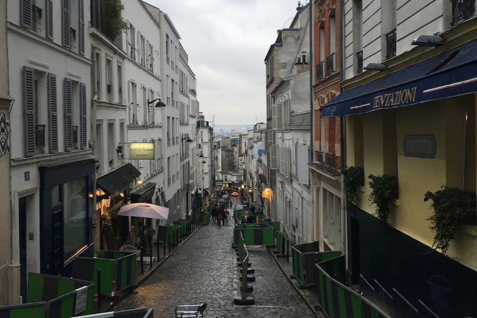 Une rue pavée bordée de bâtiments historiques, un scooter garé et une vue lointaine sur le paysage urbain.