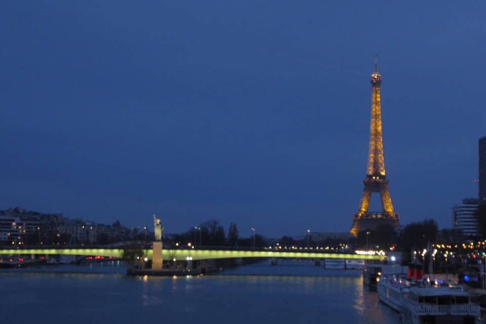 Der Eiffelturm im Abendlicht mit einer Brücke und Booten im Vordergrund.