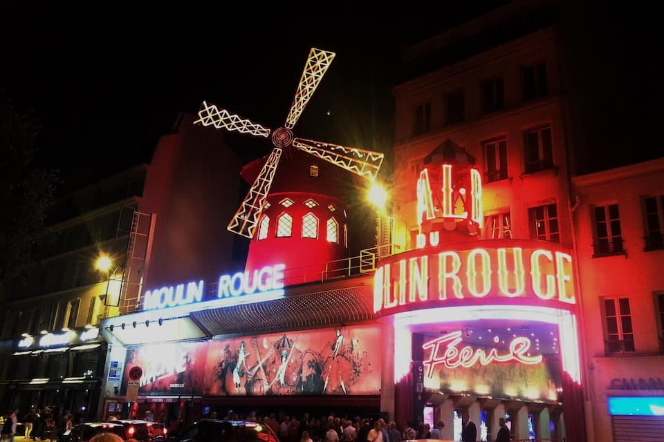 Ein hell erleuchtetes Gebäude mit Windmühle und Neonreklamen mit den Aufschriften "Moulin Rouge" und "Alain Delon".