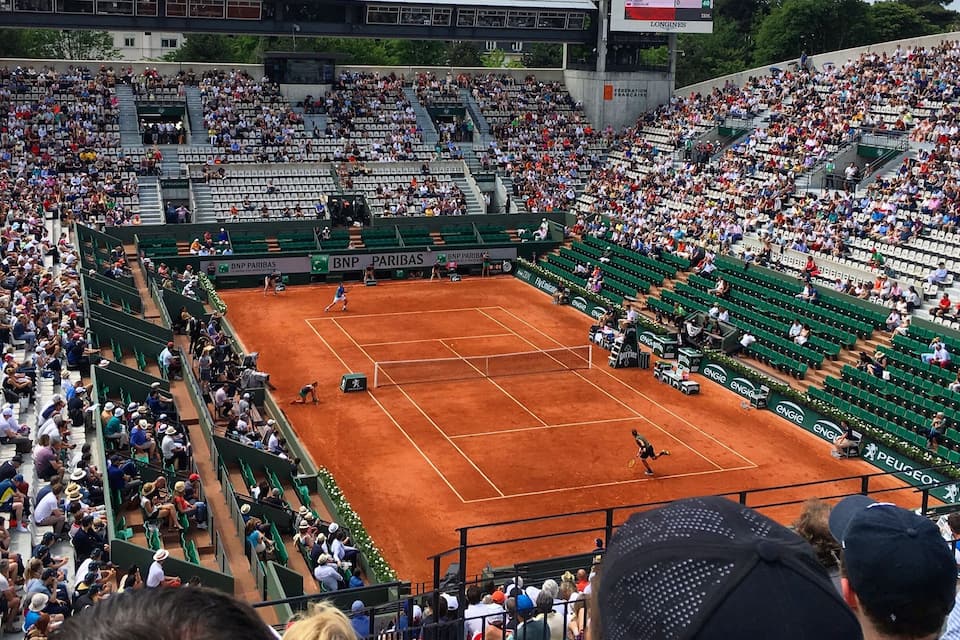 A tennis stadium with a clay court, green seating, and a scoreboard.