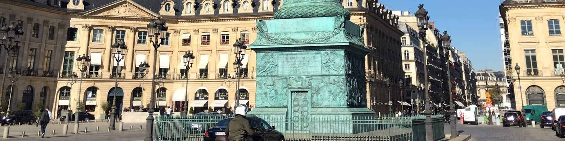 A tall, cylindrical bronze monument with intricate carvings, surrounded by historic buildings with classical architecture.