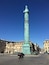 Ett högt, cylindriskt bronsmonument med detaljerade skulpturer, omgivet av historiska byggnader med klassisk arkitektur.