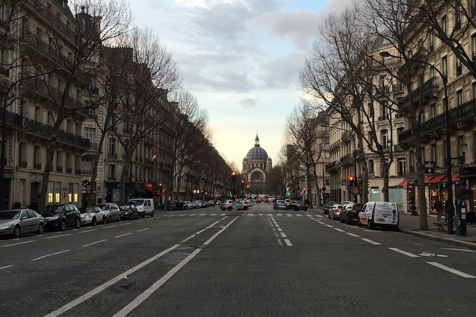 Uma rua larga com carros estacionados e edifícios em ambos os lados, conduzindo a uma grande estrutura em forma de cúpula ao longe.