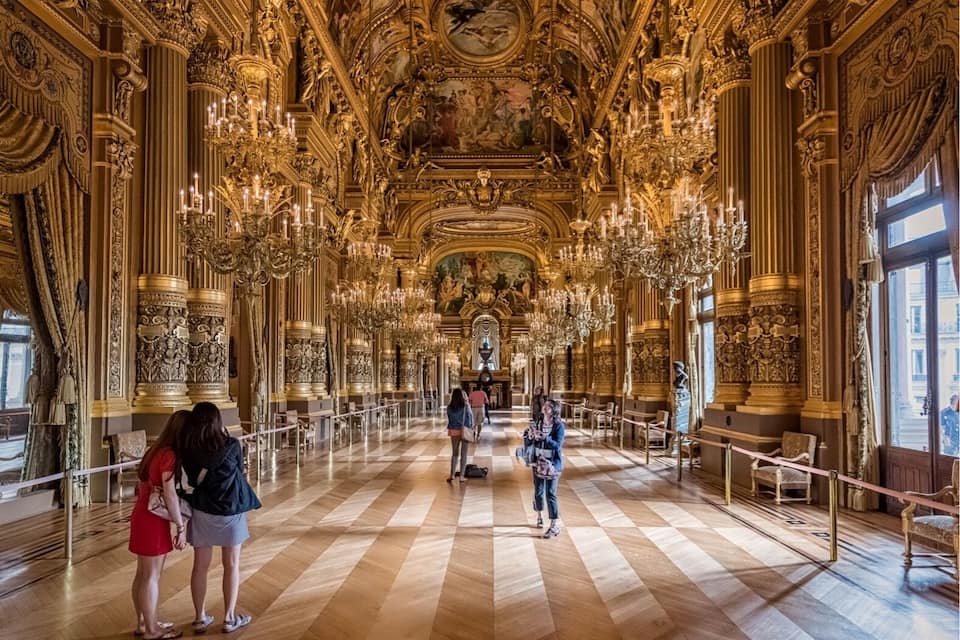 An ornate hall with intricate gold detailing, large chandeliers, and a checkered floor.