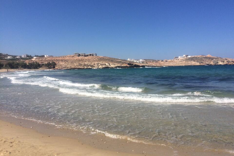 En strand med klart vand, bølger og bygninger på en klippefyldt bakke i baggrunden.