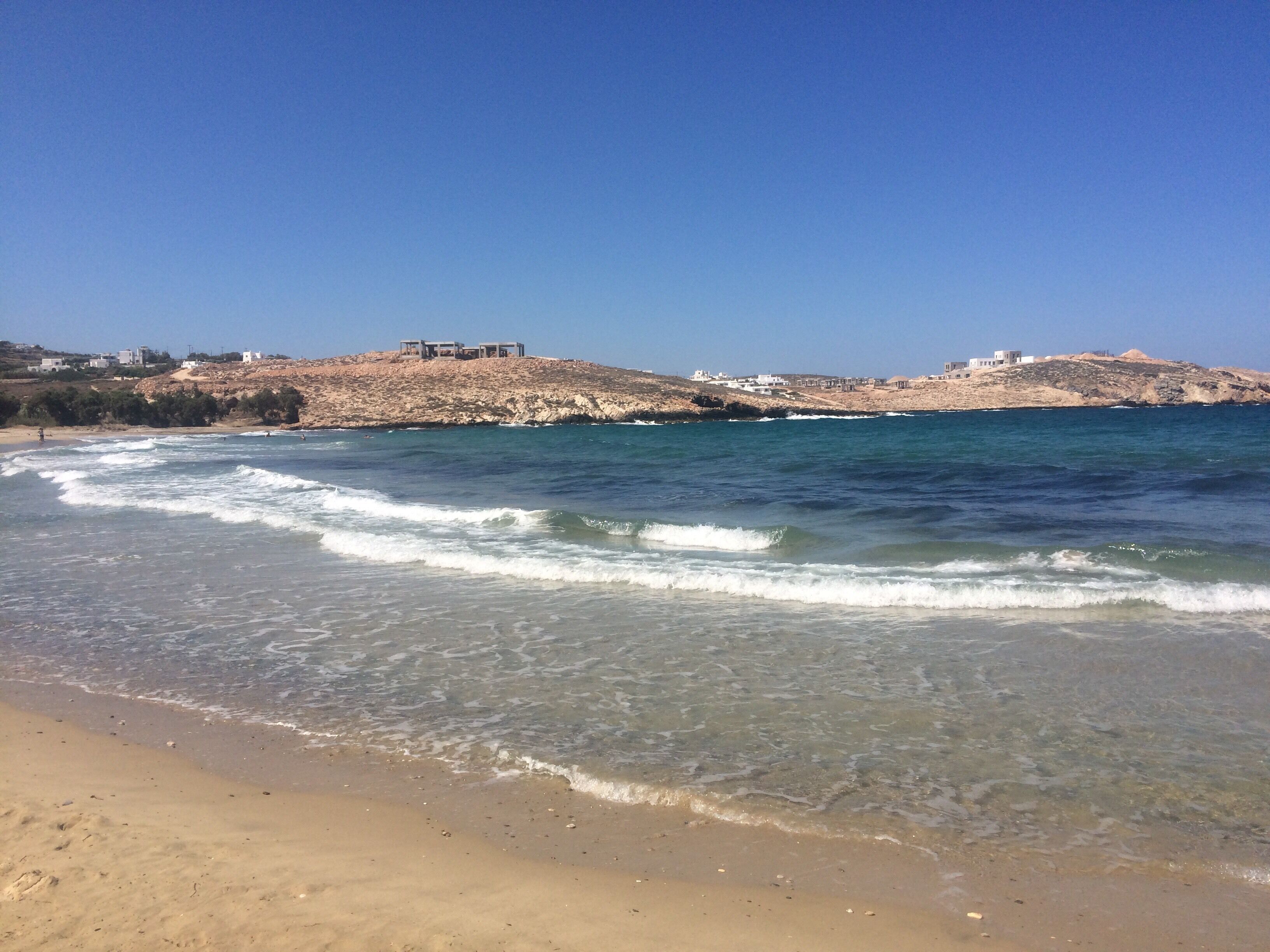 A beach with clear water, waves, and buildings on a rocky hill in the background.
