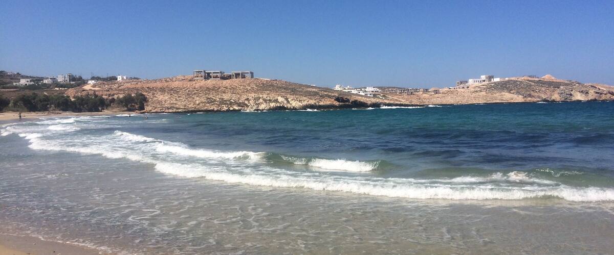 A beach with clear water, waves, and buildings on a rocky hill in the background.