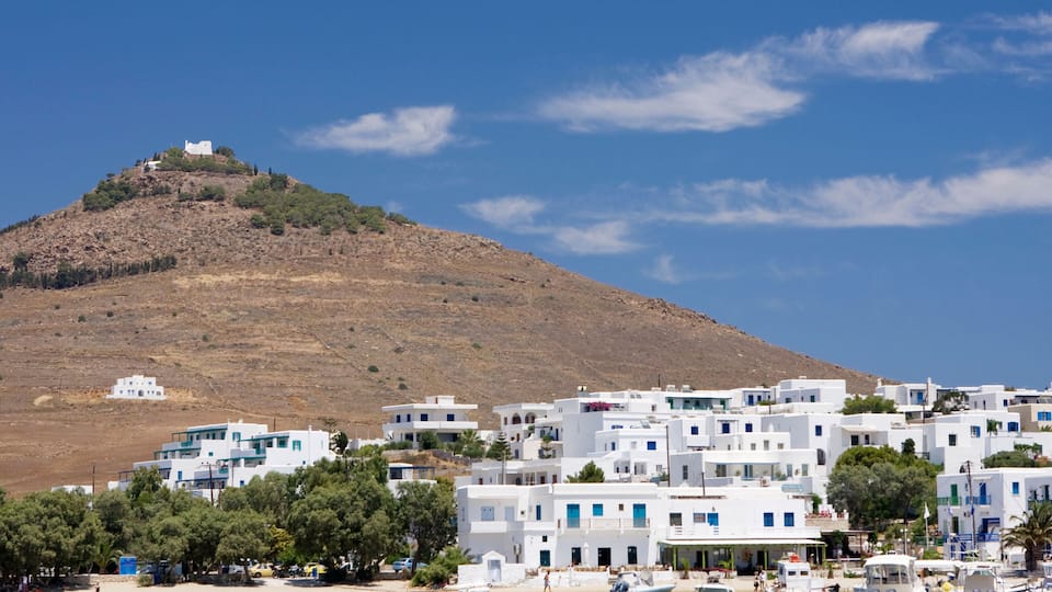 A coastal town with white buildings, a hill with a structure on top, and boats in the water.