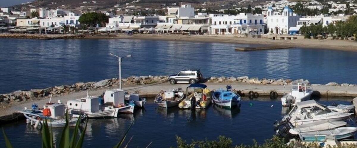 A coastal town with boats docked at a marina, surrounded by white buildings and hills in the background.