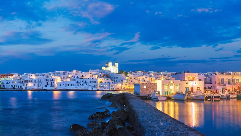 A coastal town at dusk with white buildings, a prominent church, and boats docked along the waterfront.