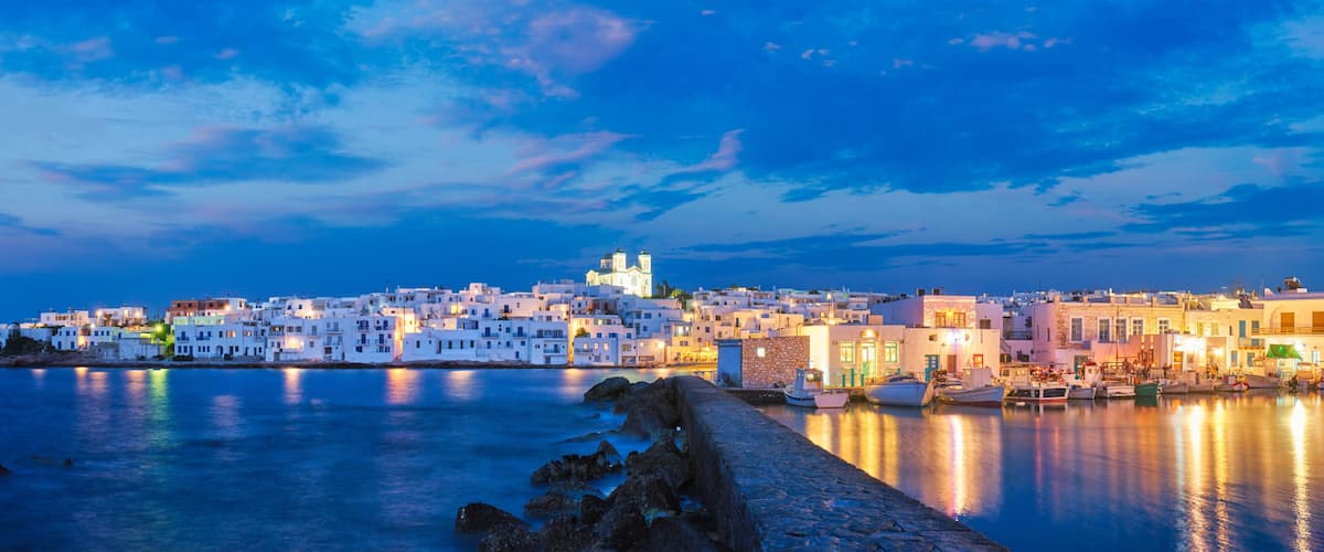 A coastal town at dusk with white buildings, a prominent church, and boats docked along the waterfront.