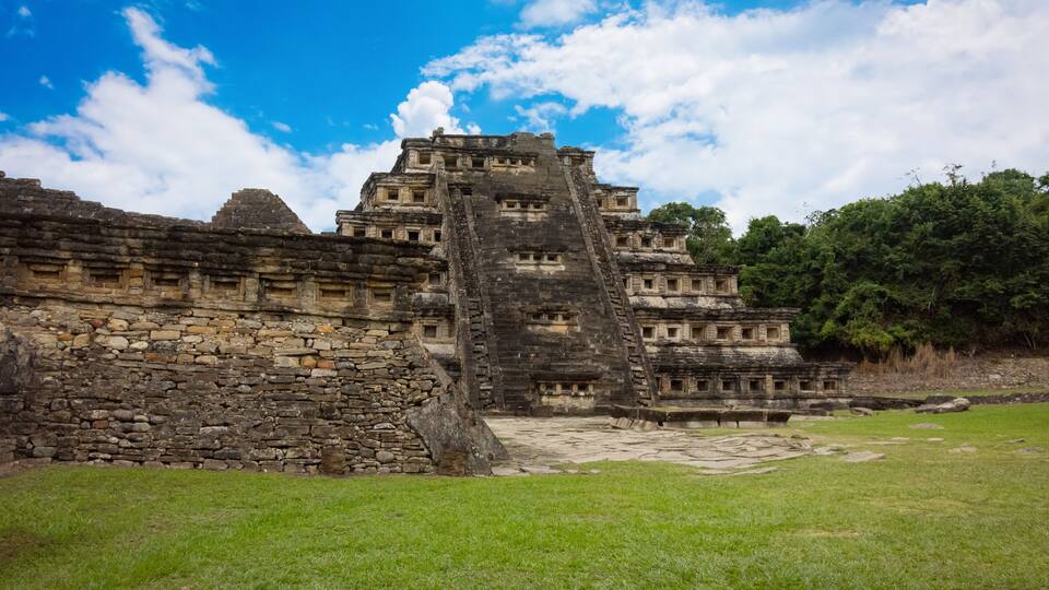 An ancient pyramid structure with a staircase leading to the top, surrounded by stone walls and a grassy area.