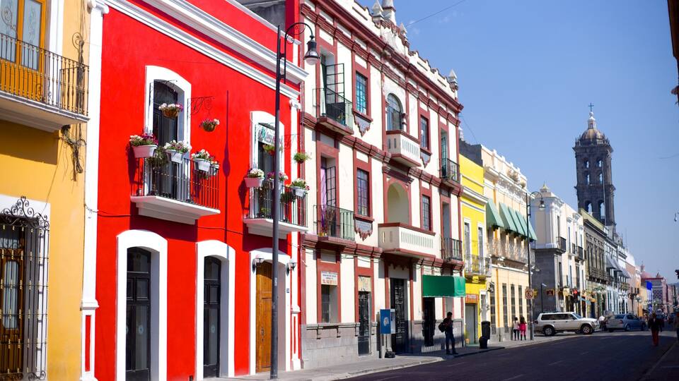 A colorful street with buildings painted in red, yellow, and white. There are balconies with flower pots, a church tower, and a street with cars and pedestrians.