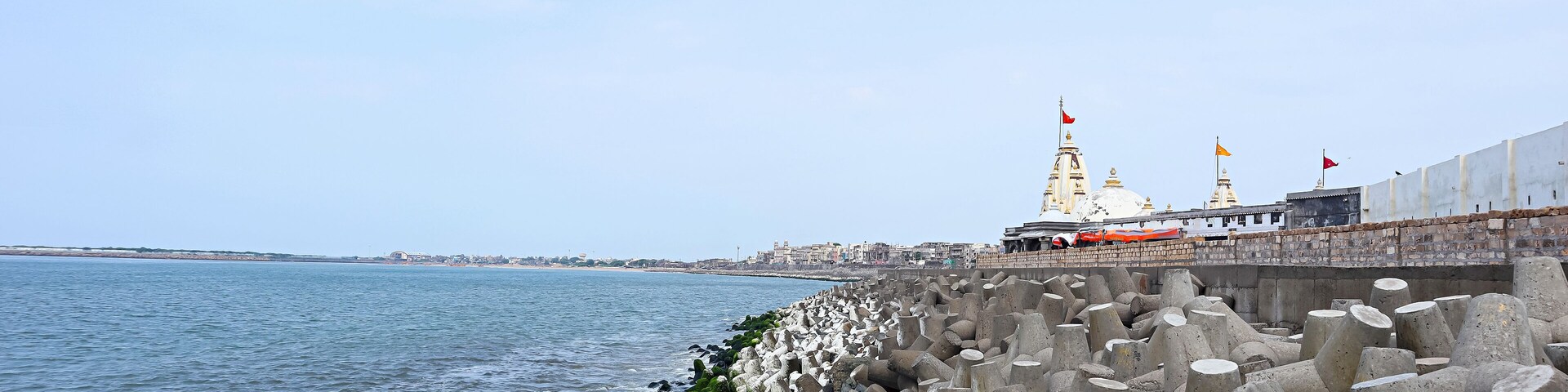 Vista costera con un muro de contención, un templo con banderas y un horizonte urbano distante.
