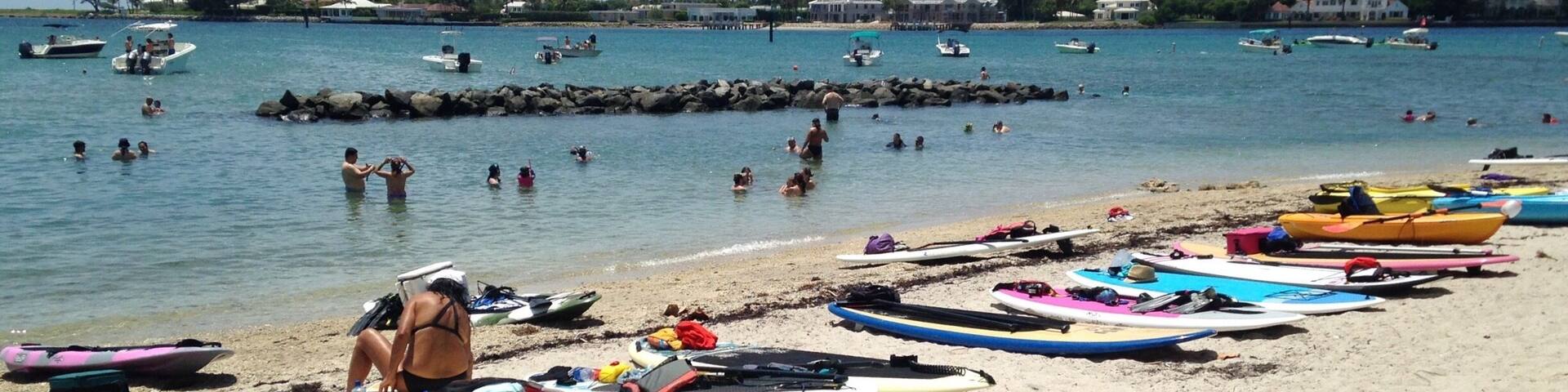 Una playa con kayaks y tablas de surf, gente nadando y barcos en el agua.