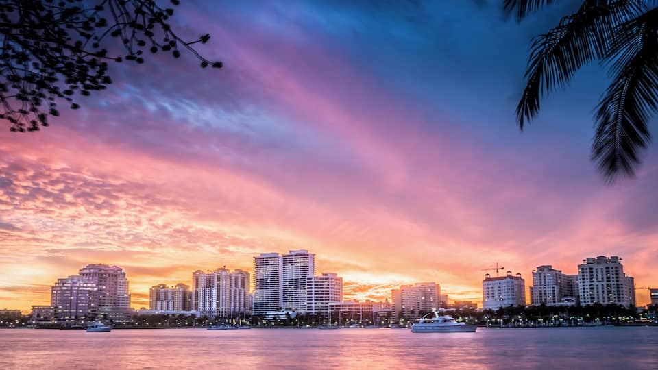 A city skyline at sunset with high-rise buildings and a body of water reflecting the sky.