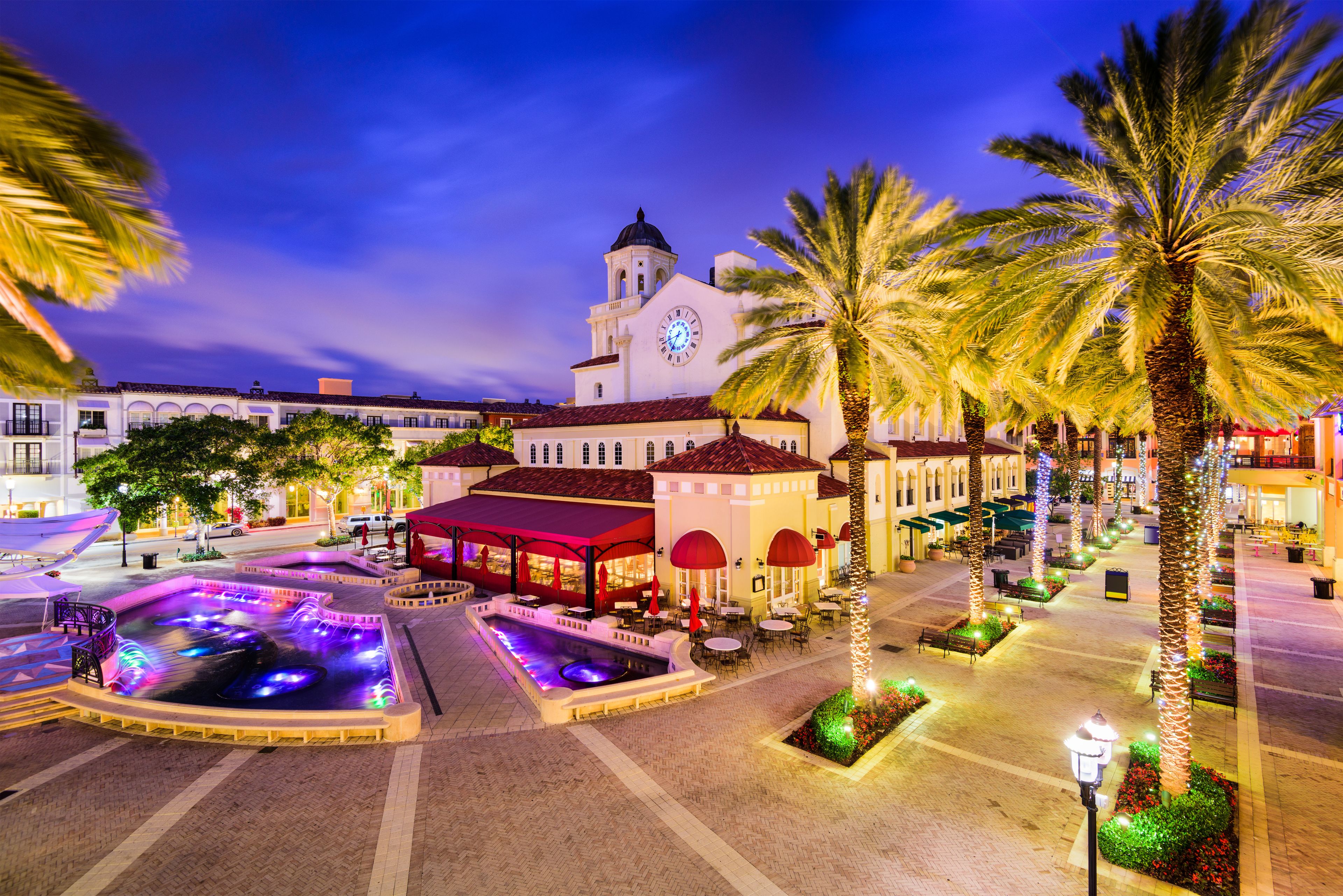 A plaza with a fountain, palm trees, and a building with a clock tower.