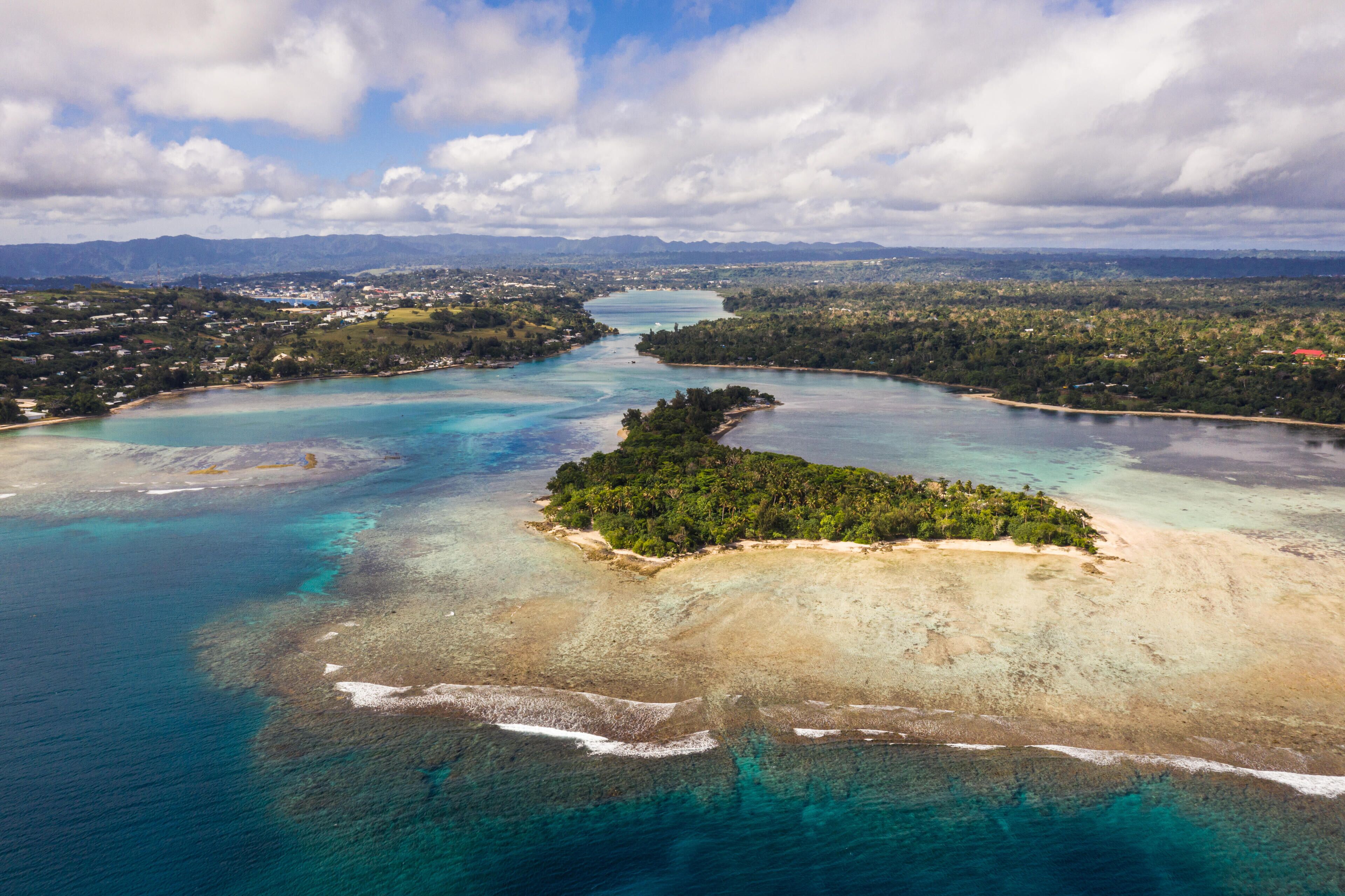 An aerial view of a coastal area with a clear blue sea, a small island with dense vegetation, and a distant cityscape.