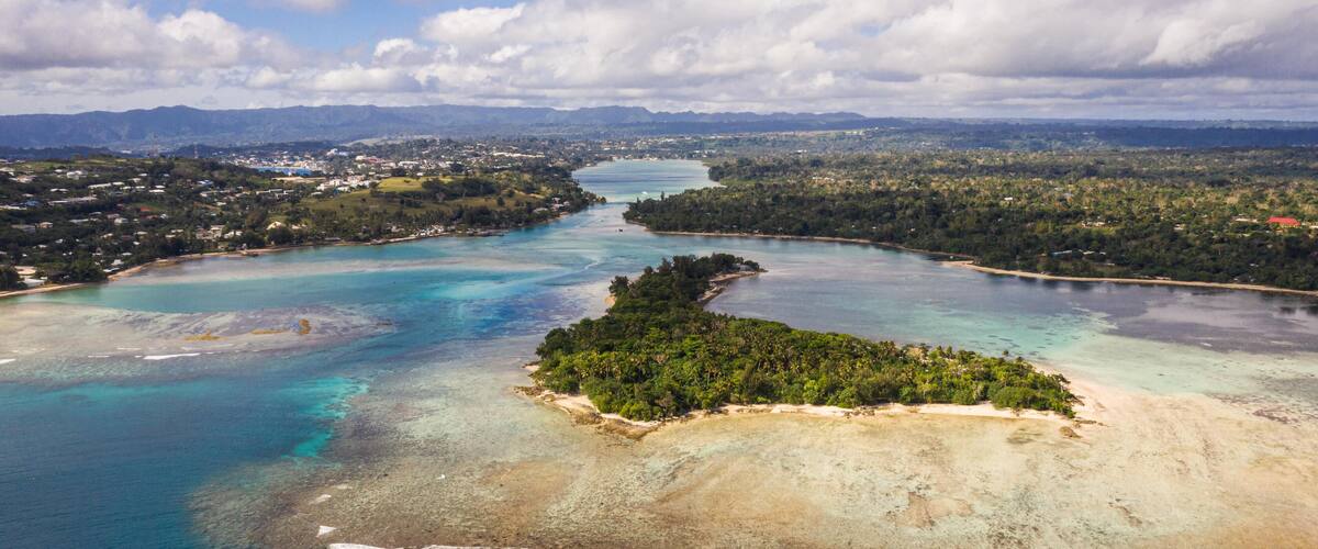 An aerial view of a coastal area with a clear blue sea, a small island with dense vegetation, and a distant cityscape.