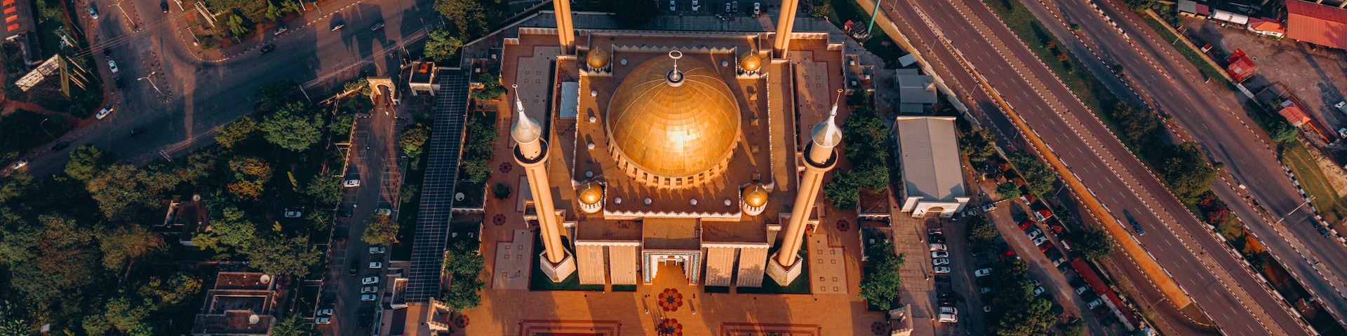 Vista aérea de uma mesquita com uma grande cúpula dourada e quatro minaretes, rodeada por estradas e vegetação.