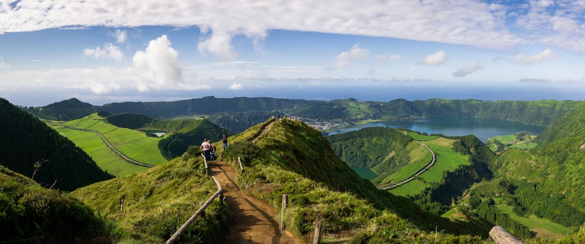 A mountainous landscape with a winding path, lush green fields, and a lake in the distance.