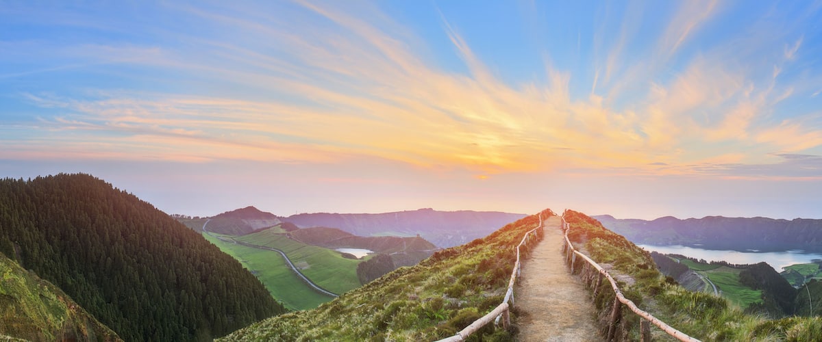 A mountain trail with a view of a valley and a lake at sunset.