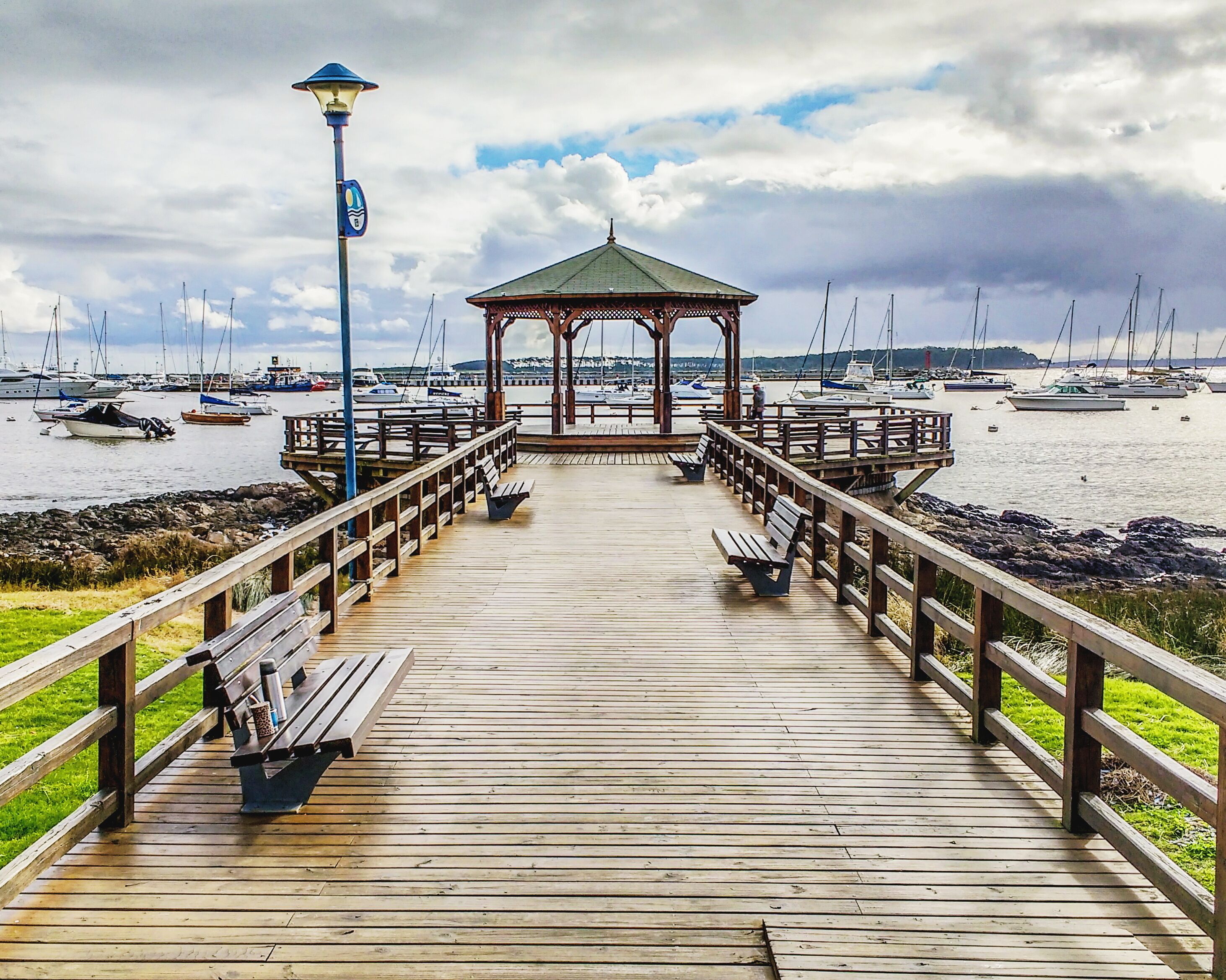 A wooden pier leading to a gazebo with benches, overlooking a marina with boats.