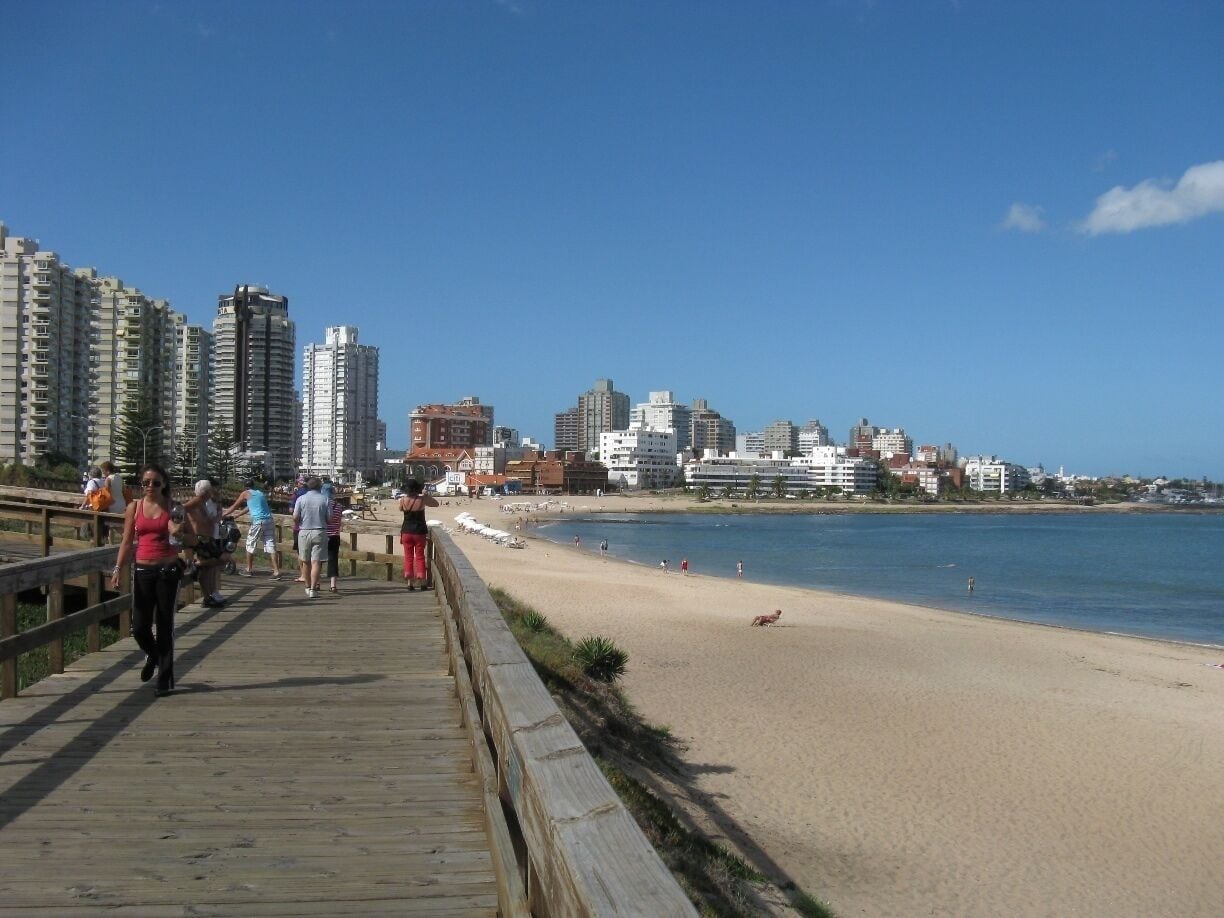 Una playa con un paseo marítimo de madera, gente caminando y edificios al fondo.