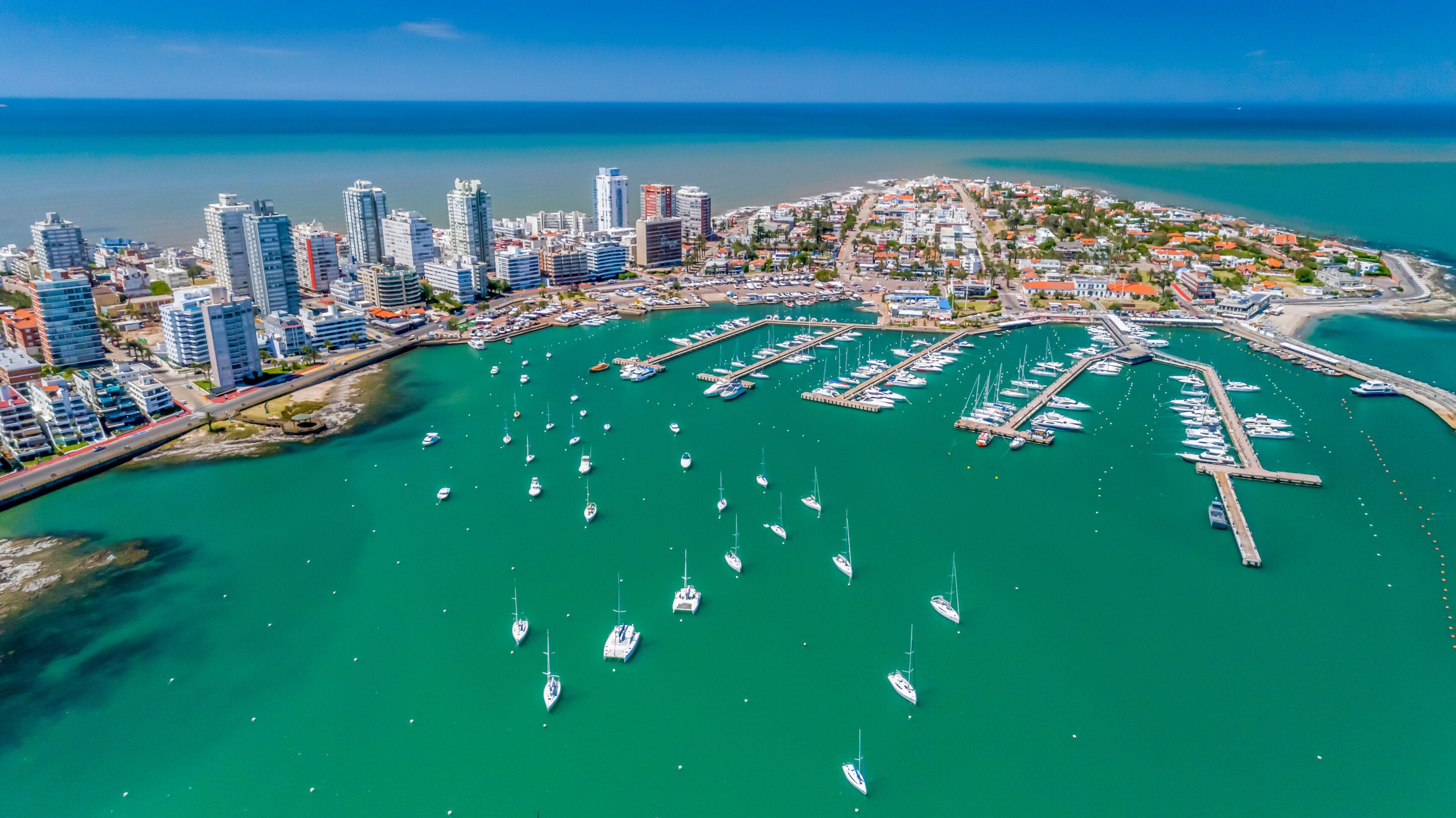 Un paisaje urbano costero con un puerto deportivo lleno de barcos, edificios altos y un mar azul cristalino.
