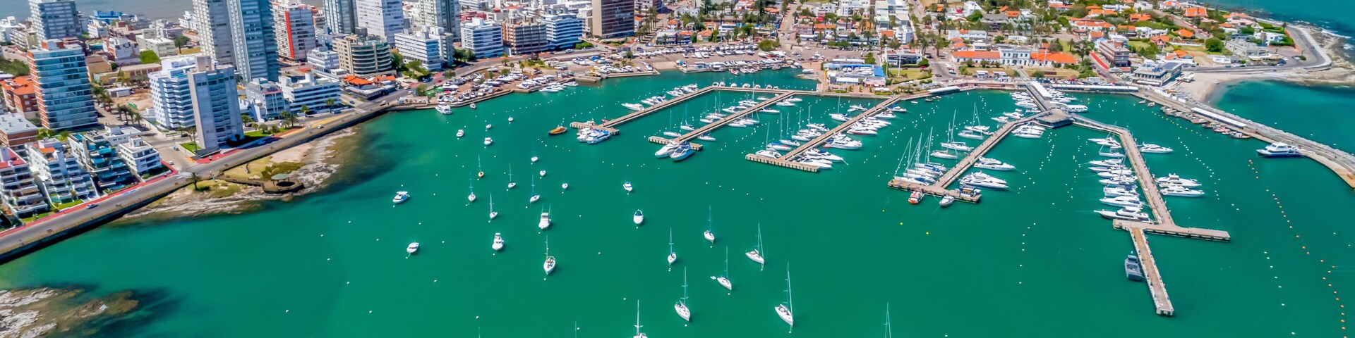 Un paisaje urbano costero con un puerto deportivo lleno de barcos, edificios altos y un mar azul cristalino.