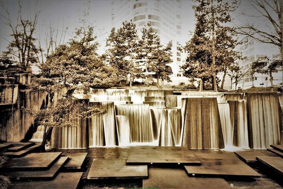 Een waterval die over houten treden stroomt, omringd door bomen en een modern gebouw op de achtergrond.