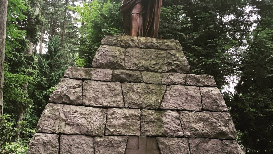 A statue of a figure with arms raised, standing on a stone pedestal in a wooded area.