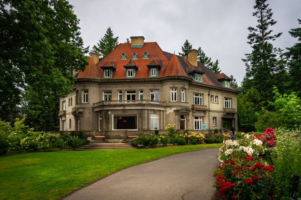 A large, historic building with a red-tiled roof and a well-maintained garden with various flowers.