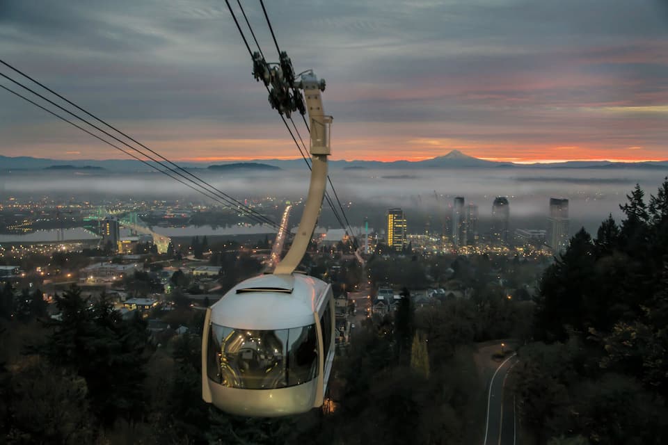 Une télécabine se déplace le long d’un câble, offrant une vue sur une ville avec une montagne en arrière-plan et un ciel au coucher du soleil.