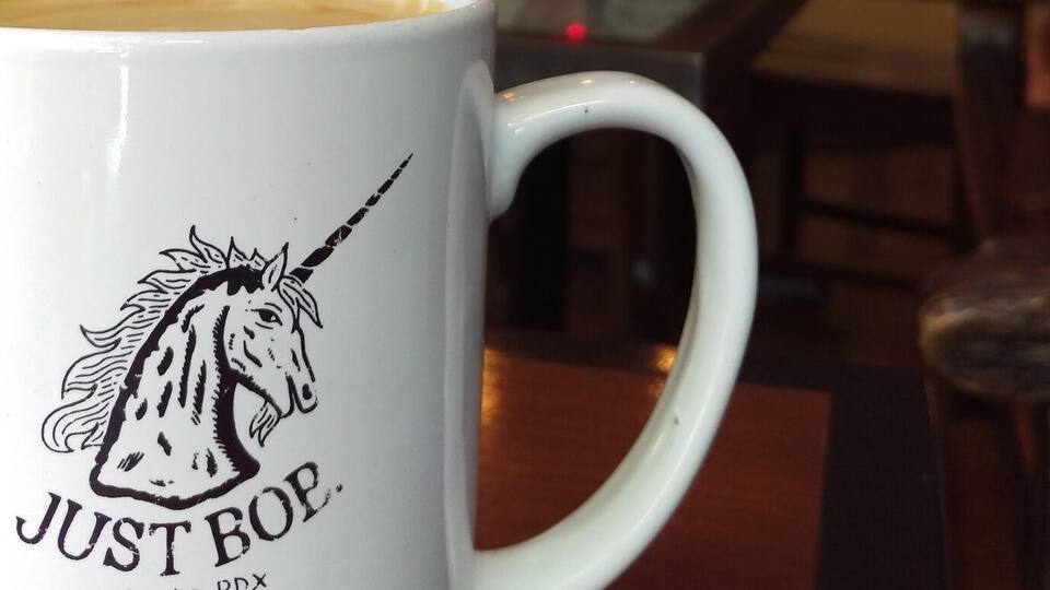 A white mug with a unicorn design and the text "JUST BOP Alberta.PPX" in a cafe setting.