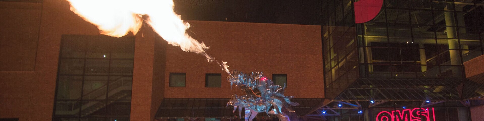 Un dragón escupiendo fuego actuando frente a un edificio con fachada de vidrio y un letrero de neón.